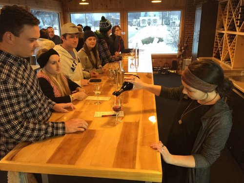 A woman pours wine at a bar while a group of people watch, enjoying a cozy indoor setting with wooden decor.