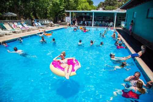 A busy pool scene with people swimming, lounging on floats, and relaxing by the water on a sunny day.