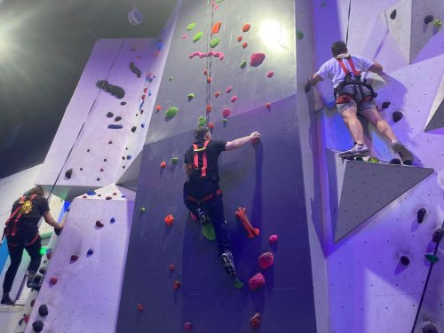 Two climbers ascend a colorful indoor rock climbing wall, with bright holds and safety harnesses.