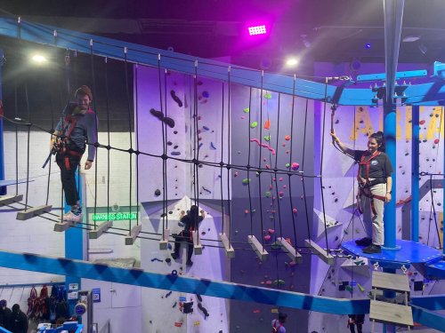 Two people navigate a rope bridge in an indoor climbing gym, surrounded by climbing walls and colorful holds.
