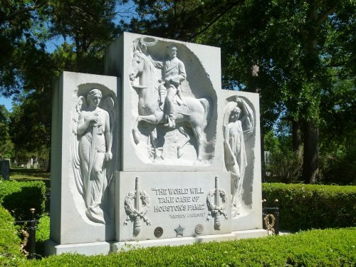 A stone monument featuring a horseman and two figures, surrounded by greenery, with an inscription about Houston's fame.