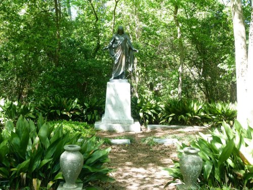 A statue of a figure in robes stands amidst lush greenery, surrounded by plants and decorative urns.