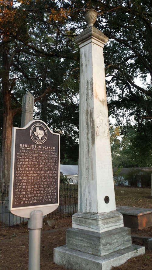 A historical marker stands next to a tall monument in a cemetery, surrounded by trees and grass.