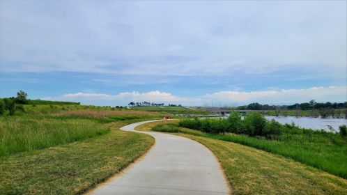 A winding path through green grass leads to a river under a blue sky with scattered clouds.
