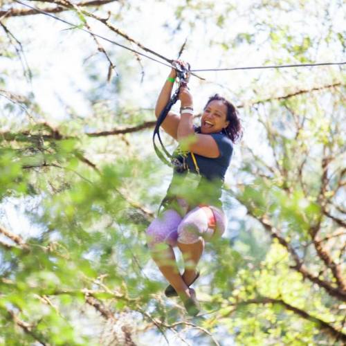 A woman joyfully zip-lining through a forest, surrounded by green trees and sunlight.