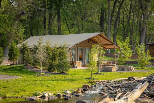 A cozy tent cabin surrounded by greenery and trees, with a small stream and picnic area nearby.