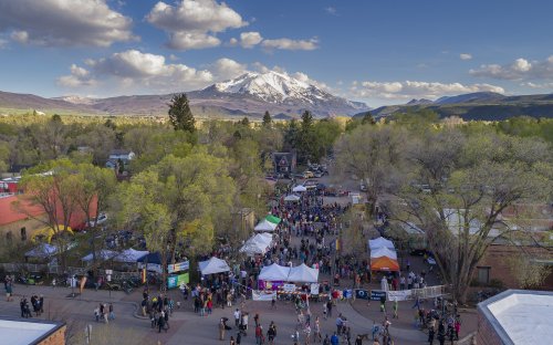 Aerial view of a bustling outdoor festival with tents, trees, and snow-capped mountains in the background.