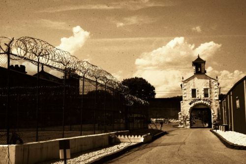 A sepia-toned image of a prison entrance with barbed wire and a watchtower under a cloudy sky.
