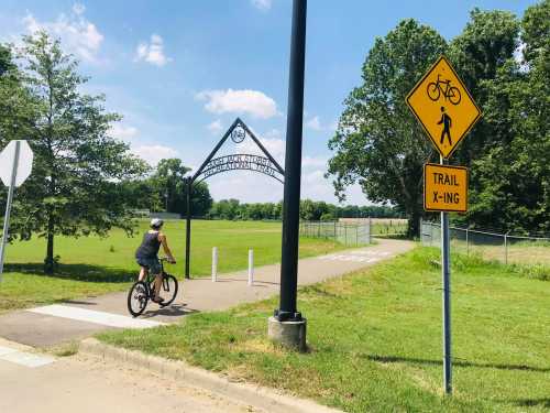 A cyclist rides on a paved trail near a sign indicating a bike and pedestrian crossing. Lush greenery surrounds the area.