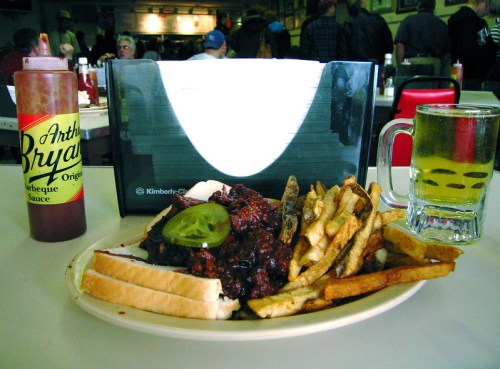 A plate of barbecue sandwich with pickles, fries, and a beer, alongside a bottle of barbecue sauce on a table.