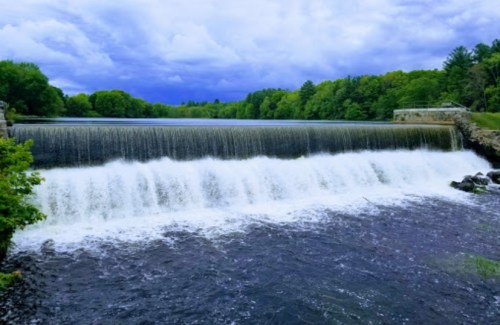 A serene waterfall cascades over a dam, surrounded by lush green trees under a cloudy sky.