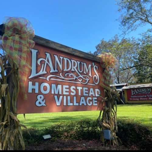 Sign for Landrum's Homestead & Village, decorated with colorful bows and surrounded by greenery under a clear blue sky.