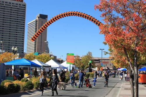 A lively street scene with people walking, colorful tents, and an arch made of orange balloons against a city backdrop.
