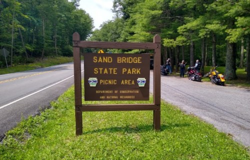 Sign for Sand Bridge State Park Picnic Area, surrounded by trees, with a road and parked motorcycles nearby.