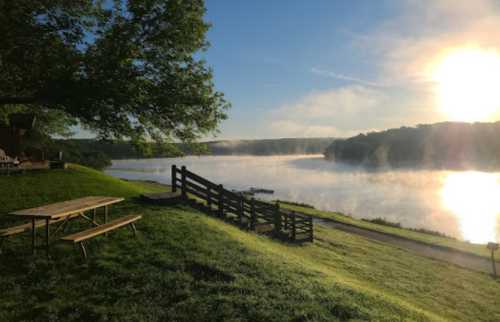 A serene lakeside view at sunrise, with mist rising over the water and a grassy area with a picnic table.