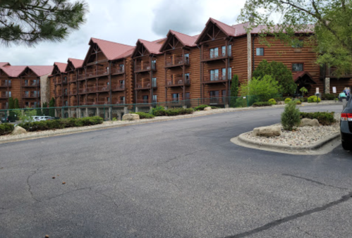 A large log cabin-style building with a red roof, surrounded by greenery and a paved parking area.