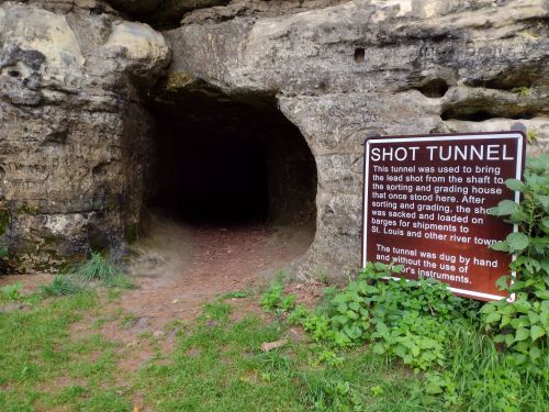 A dark tunnel entrance in a rocky landscape, with a sign explaining its historical use for transporting shot.