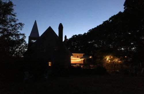 A silhouette of a house at dusk, with string lights illuminating a deck and trees in the background.