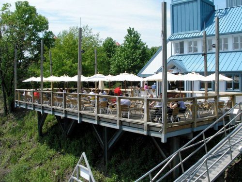 A wooden deck with tables and umbrellas, overlooking greenery, attached to a building with a blue roof.