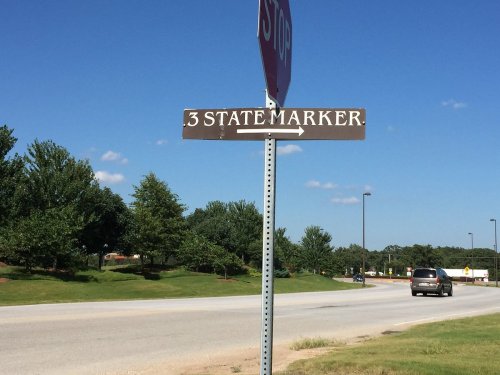 A brown sign indicating "3 State Marker" with arrows pointing left and right, near a stop sign and grassy area.