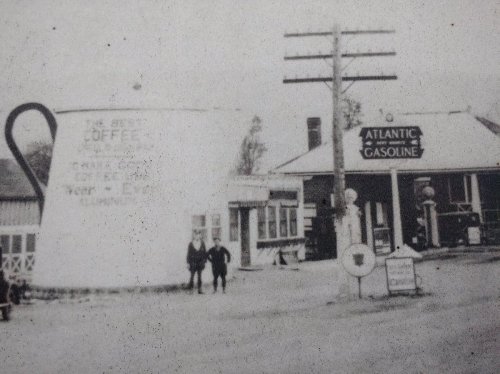 Historic black-and-white photo of a large coffee cup sign and a gas station, with two people standing in front.