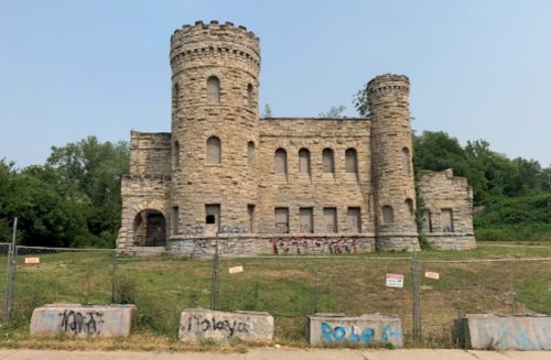 Abandoned stone castle with two towers, surrounded by greenery and covered in graffiti. Fenced off for safety.