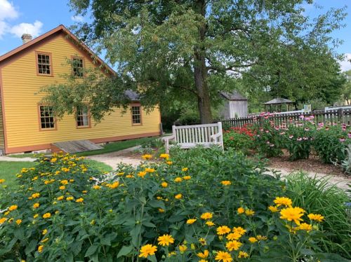 A yellow house surrounded by vibrant flowers and greenery, with a wooden bench in a peaceful garden setting.