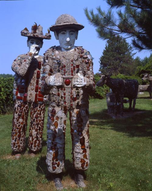 Two whimsical stone sculptures of men, adorned with colorful glass, stand in a grassy area with trees and a cow statue nearby.