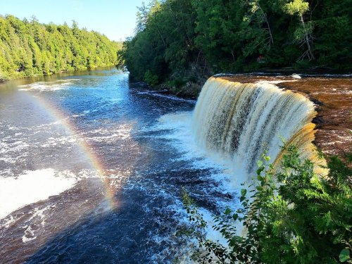 A scenic waterfall cascading into a river, surrounded by lush green trees and a faint rainbow in the mist.