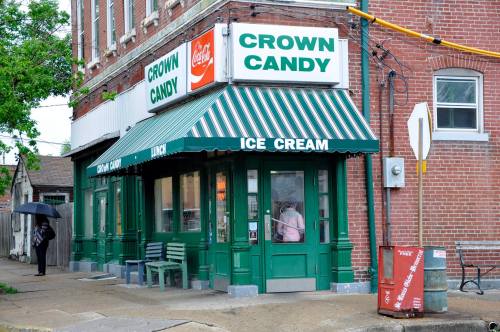 A quaint ice cream shop with a green and white striped awning, featuring "Crown Candy" signage and a person under an umbrella.