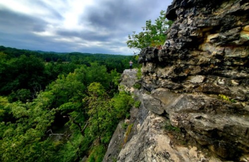 A person stands on a rocky cliff overlooking a lush green landscape under a cloudy sky.