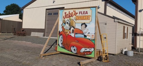 A colorful sign for "Jake's Flea Market" featuring a cartoon car and characters, set against a rustic building backdrop.