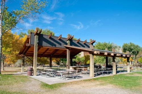 A spacious picnic shelter with wooden beams and tables, surrounded by trees and a clear blue sky.