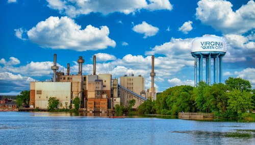 Industrial buildings beside a river, with a water tower labeled "Virginia Queen City" and a blue sky with clouds.