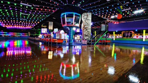 A brightly lit indoor skating rink with colorful lights, arcade games, and a polished wooden floor reflecting the lights.