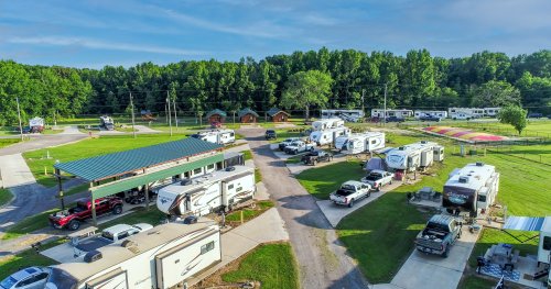 Aerial view of a campground with RVs, cars, and cabins surrounded by green trees and open fields.