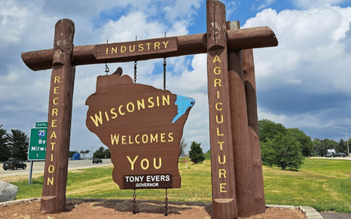 Wooden welcome sign for Wisconsin featuring the state outline and text: "Wisconsin Welcomes You" with "Tony Evers."