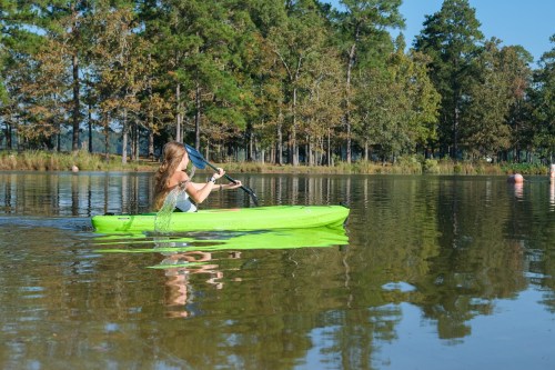 A person paddles a bright green kayak on a calm lake surrounded by trees under a clear blue sky.