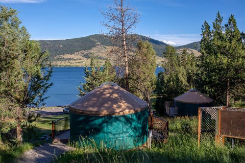 Two green yurts nestled among trees by a serene lake, with mountains in the background under a clear blue sky.