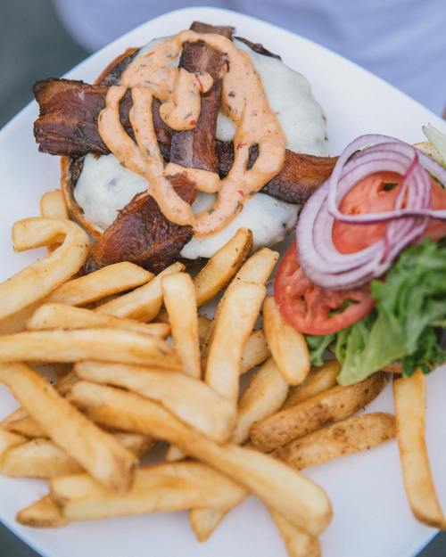 A plate with a burger topped with sauce, bacon, and cheese, accompanied by fries, lettuce, tomato, and onion.