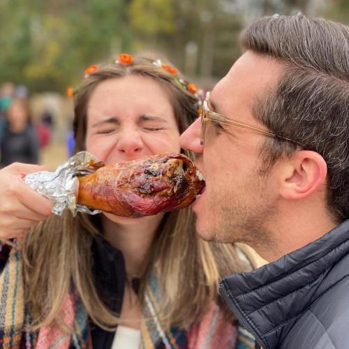 A woman playfully feeding a man a large turkey leg, both smiling and enjoying a festive outdoor setting.