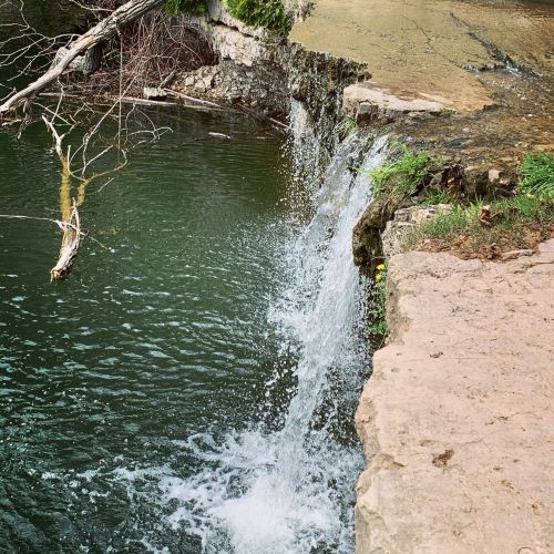 A small waterfall cascading into a serene green pool, surrounded by rocks and lush vegetation.