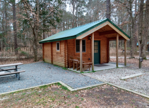 A small wooden cabin with a green roof, surrounded by trees and a gravel area with a picnic table.