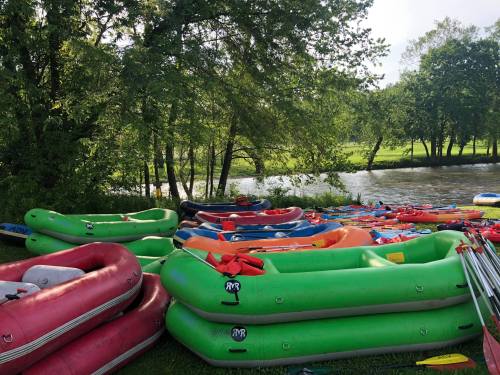 Colorful inflatable rafts stacked by a river, surrounded by lush green trees and grass.