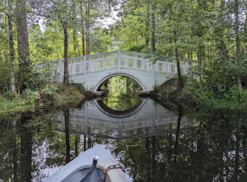 A serene view of a white bridge arching over a calm, reflective waterway surrounded by lush greenery.