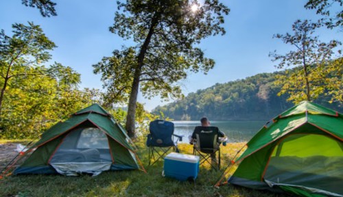 A serene campsite by a lake, featuring two green tents and a person sitting in a chair, surrounded by trees.