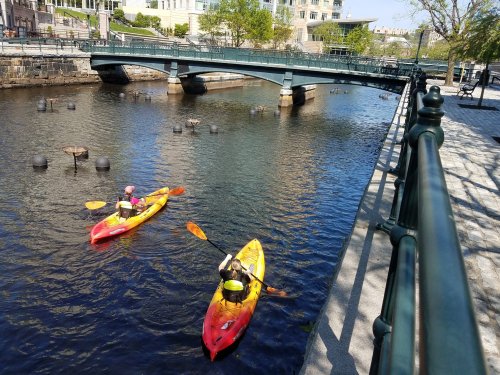 Two kayakers paddle in a calm river beneath a bridge, surrounded by greenery and urban buildings.
