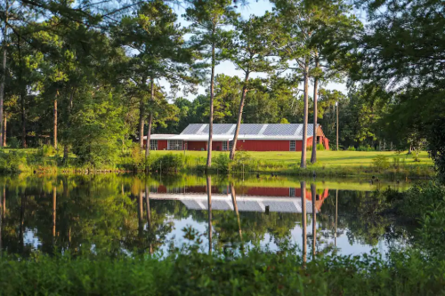 A red barn-style building surrounded by trees, reflecting in a calm pond under a clear blue sky.