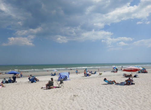 A sunny beach scene with people relaxing on sand, colorful umbrellas, and gentle waves in the background.