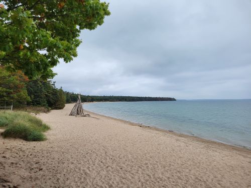 A sandy beach with a wooden structure, surrounded by trees and calm water under a cloudy sky.